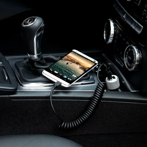 Silverfärgad USB-billaddare med två portar och spiralkabel, ansluten i bilens 12V-uttag, laddar smartphone i bilen.