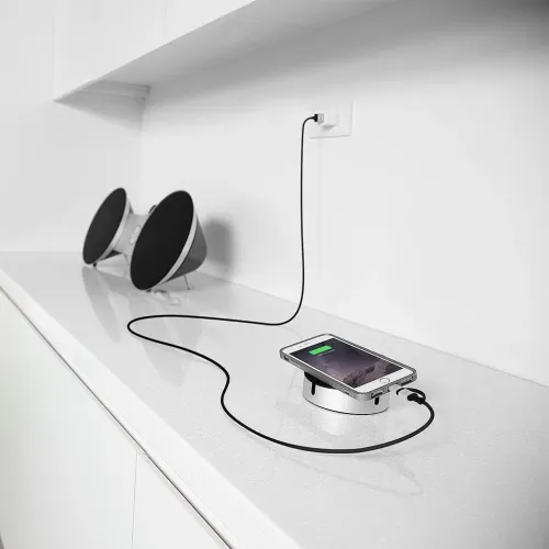 Langes Ladekabel mit USB-Stecker in einem Steckdosenadapter, verläuft über eine weiße Arbeitsplatte und ist mit einem Smartphone auf einer runden Aluminium-Dockingstation verbunden.