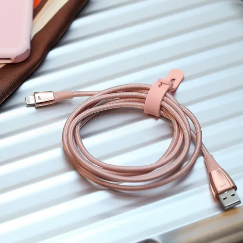 Roséguld Lightning-kabel med USB‑A och Lightning-kontakter, flätad och hoprullad med kabelrem på räfflad metallpanel.