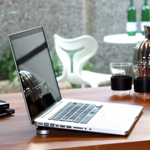 Lazy Couch aluminiumstativ lyfter bakkanten på en laptop; rund puck under vänster hörn med låg profil, placerat på träbord.