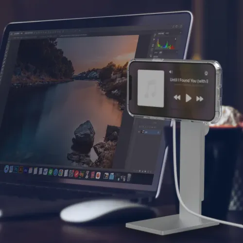 The AluDisc Pro aluminum iPhone stand holds a landscape iPhone with attached cable next to a laptop on a desk.