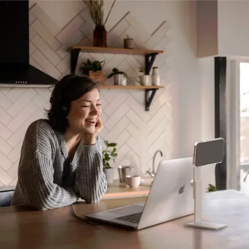 iPhone stand in white aluminum with rectangular base and narrow column, on table next to laptop, holds phone in portrait position.